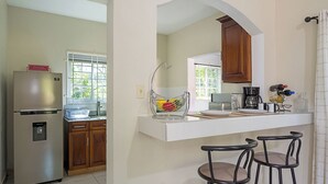 A kitchen with a stainless steel refrigerator, wooden cabinets, a white countertop, a hanging fruit basket, and two bar stools.