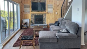 A spacious living room with a large grey sofa, a flat-screen TV mounted above a stone fireplace, and a wooden staircase.
