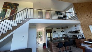 A spacious living area with a high ceiling, a staircase leading to a balcony, and a modern kitchen with an island.