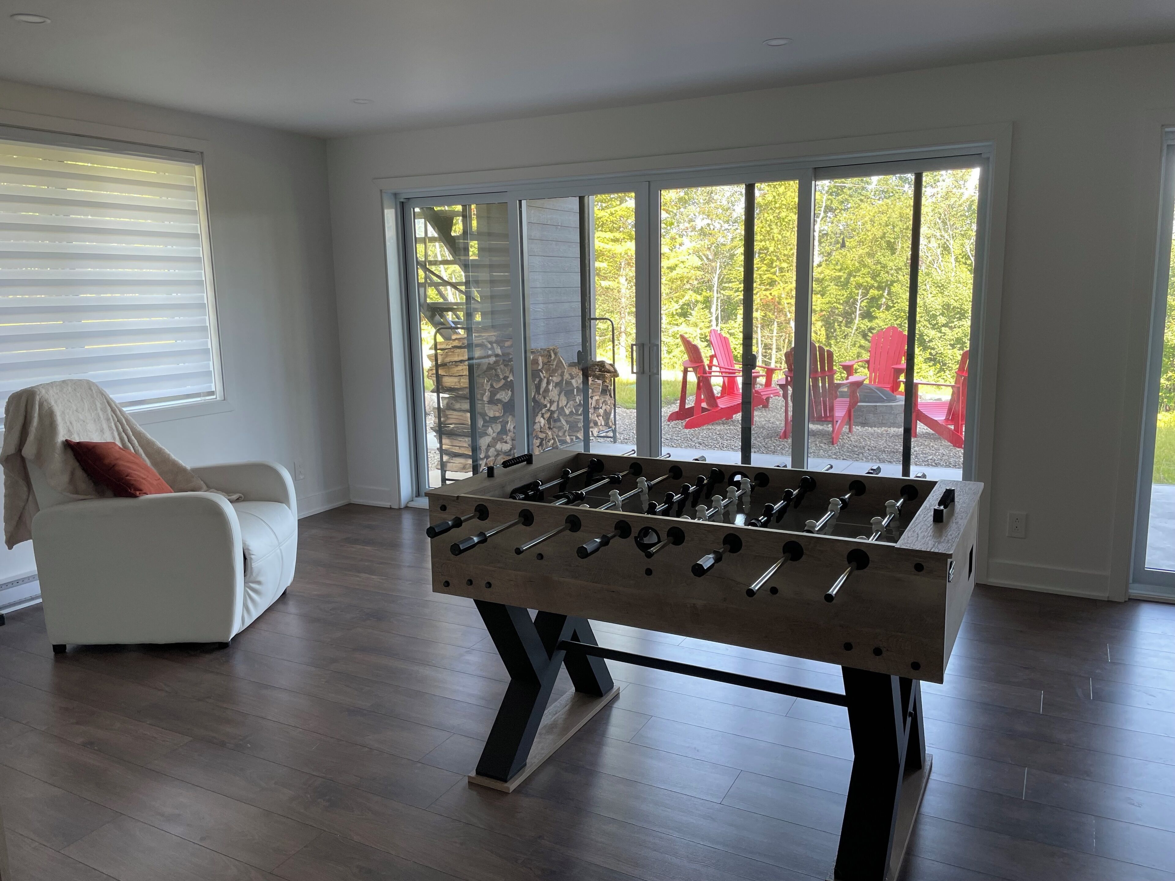 Una sala de estar moderna con una mesa de fútbol de mesa, un sillón blanco y un ventanal con vistas a un área de estar exterior.