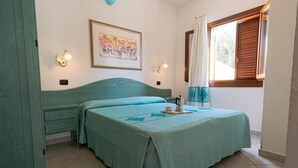 A bedroom with a bed, a nightstand, a wall-mounted lamp, a window with curtains, and a painting on the wall.