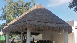 Un área de estar al aire libre cubierta con techo de paja, rodeada de vegetación y una piscina.