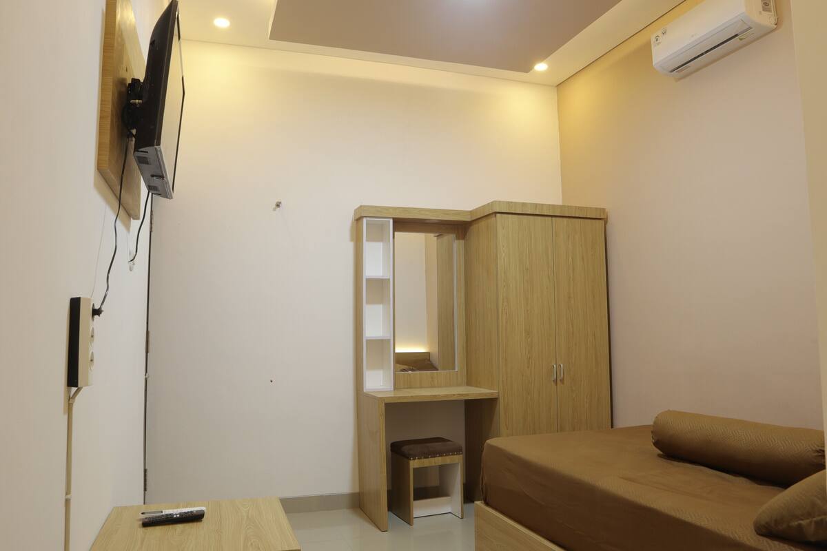 A compact bedroom with a bed, a wooden wardrobe, a small desk with a chair, and a mounted TV.