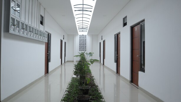 A hallway with a central plant, white walls, and doors numbered 02 to 07. - The Maguwo Premiere Kost Ekslusif (Depok)