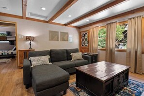 A cozy living room with a dark grey sectional sofa, a wooden coffee table, and a view of greenery through a large window with beige curtains. - Serene Scene• Spa • Close to Bear Mtn (Big Bear Lake)