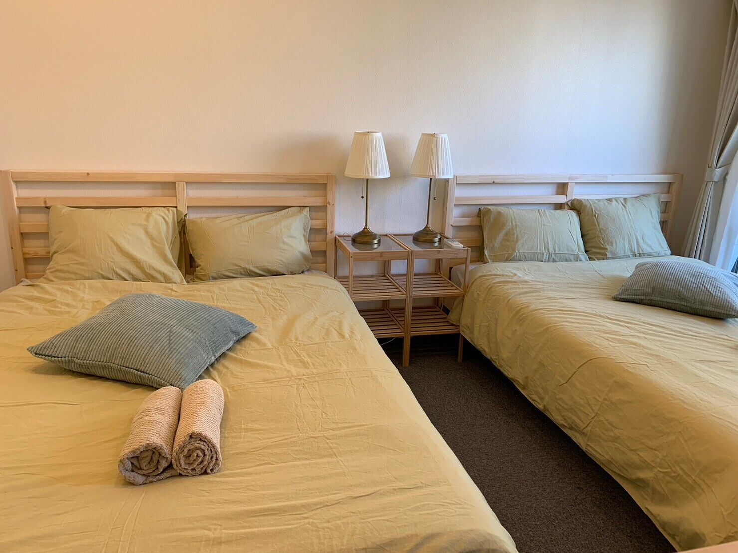 A hotel room with two beds, each with a wooden headboard, beige bedding, and matching pillows. There are two bedside lamps on a wooden nightstand between the beds.