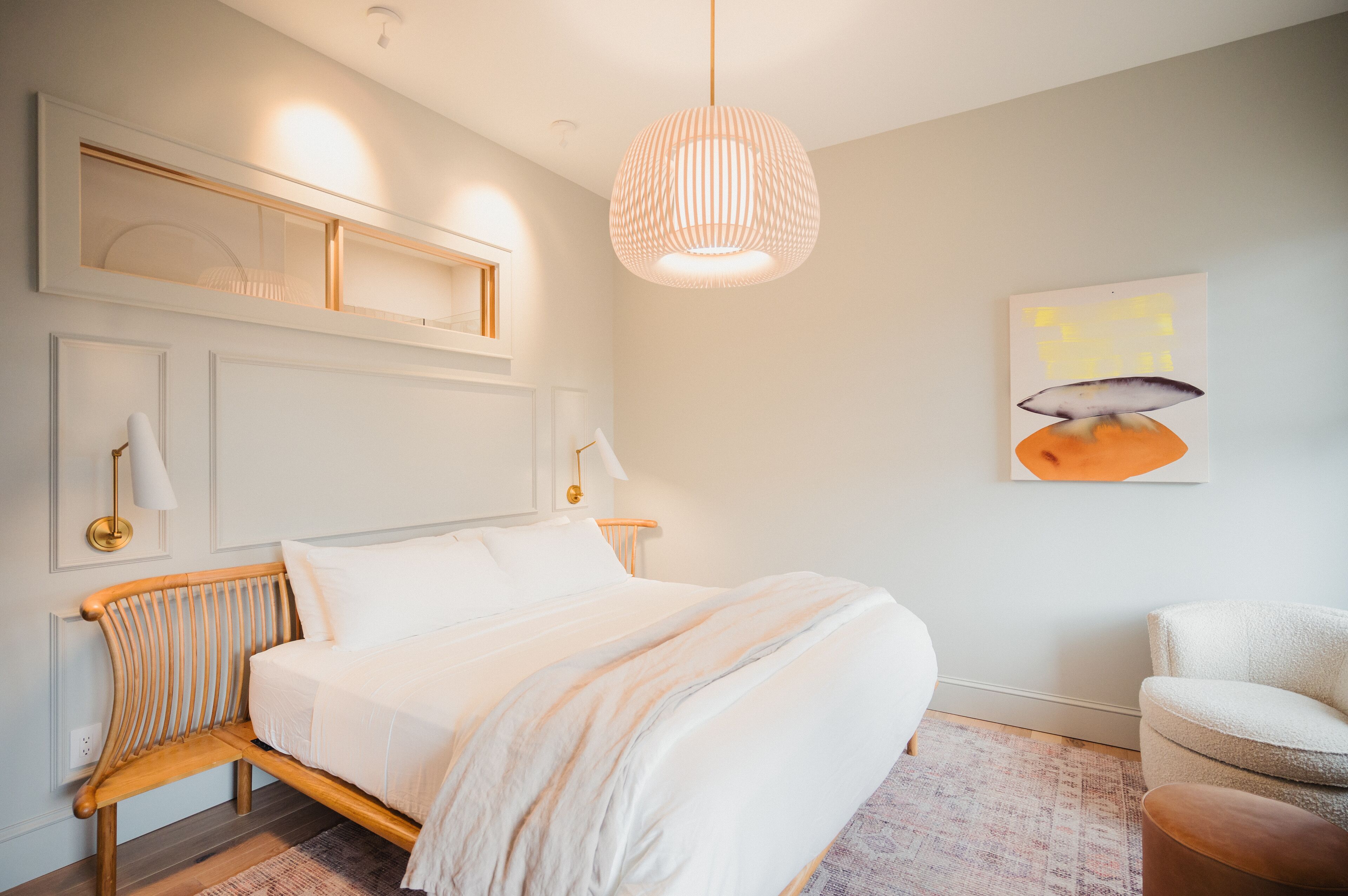 A modern bedroom with a large bed, a wooden bench, a hanging light fixture, and a wall-mounted artwork.