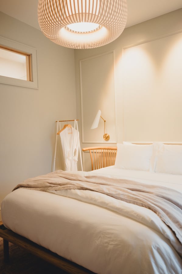 A bedroom with a bed, bedside table, lamp, and a hanging robe rack. - St. James Social (Bayfield)