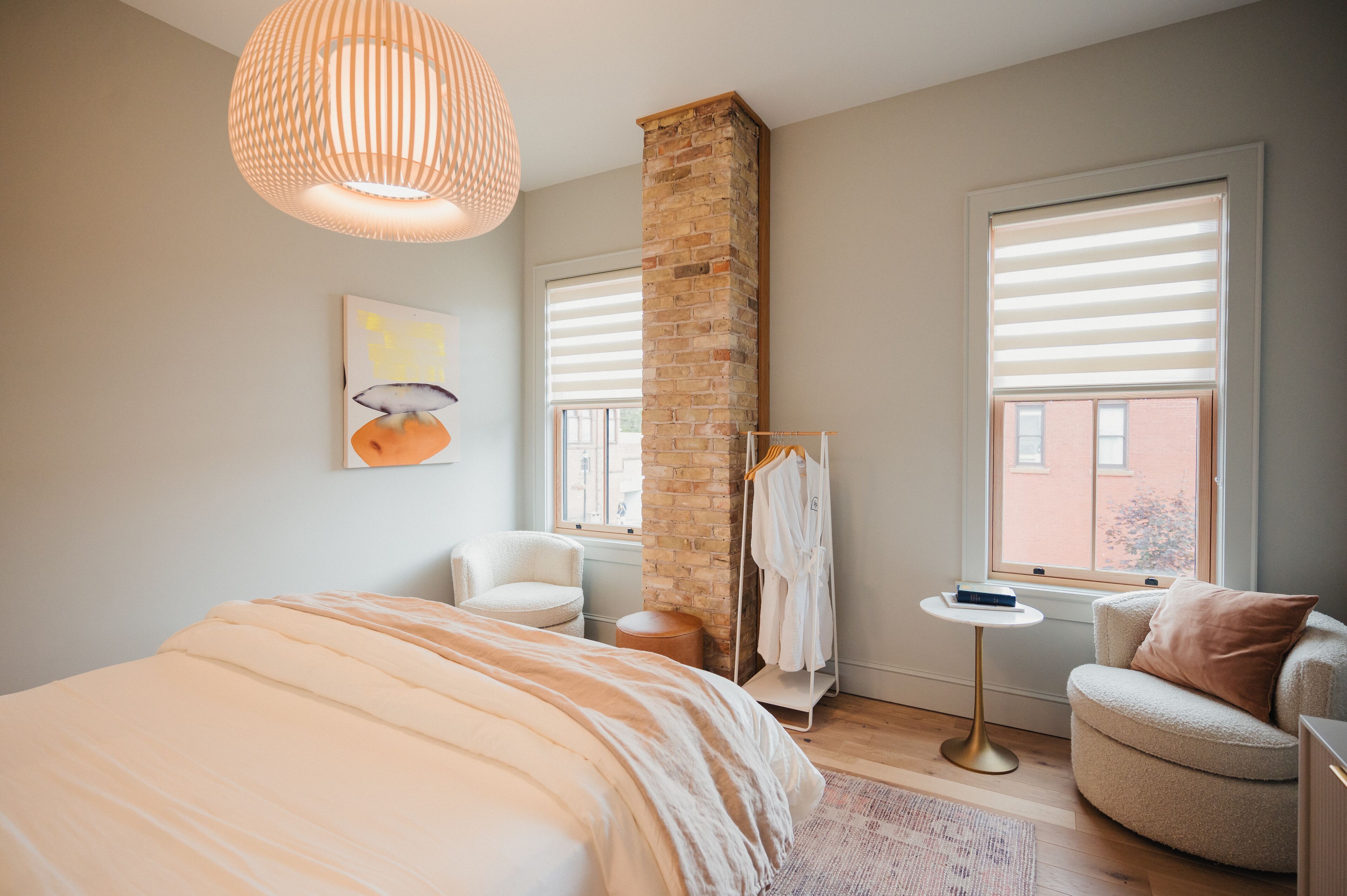 A bedroom with a large bed, a round armchair, a small table, a robe rack, and a brick column.