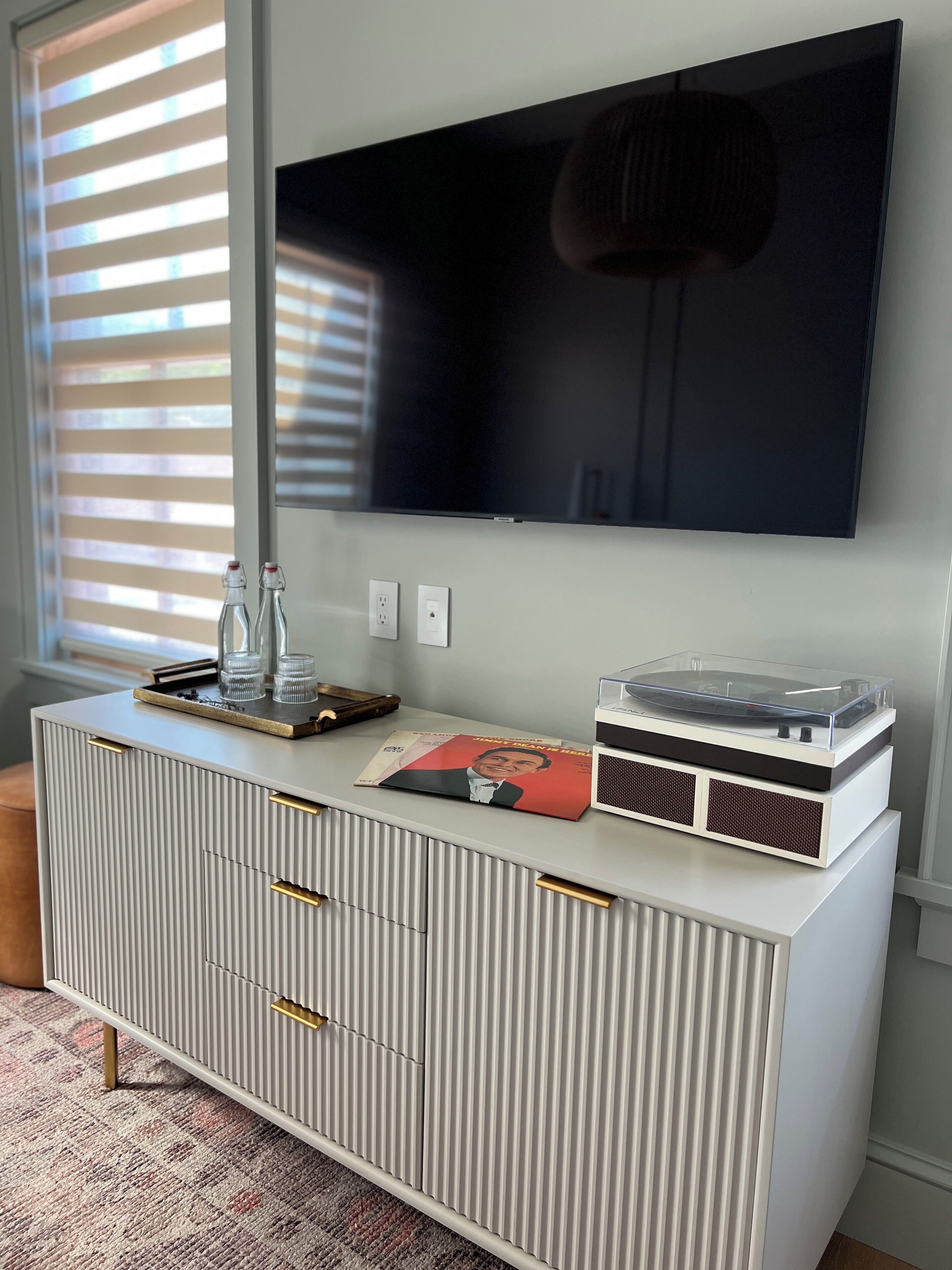 Una sala de estar moderna con un televisor de pantalla plana montado en la pared, un mueble blanco para entretenimiento con cajones, una mesa de centro de vidrio y un tocadiscos de vinilo.