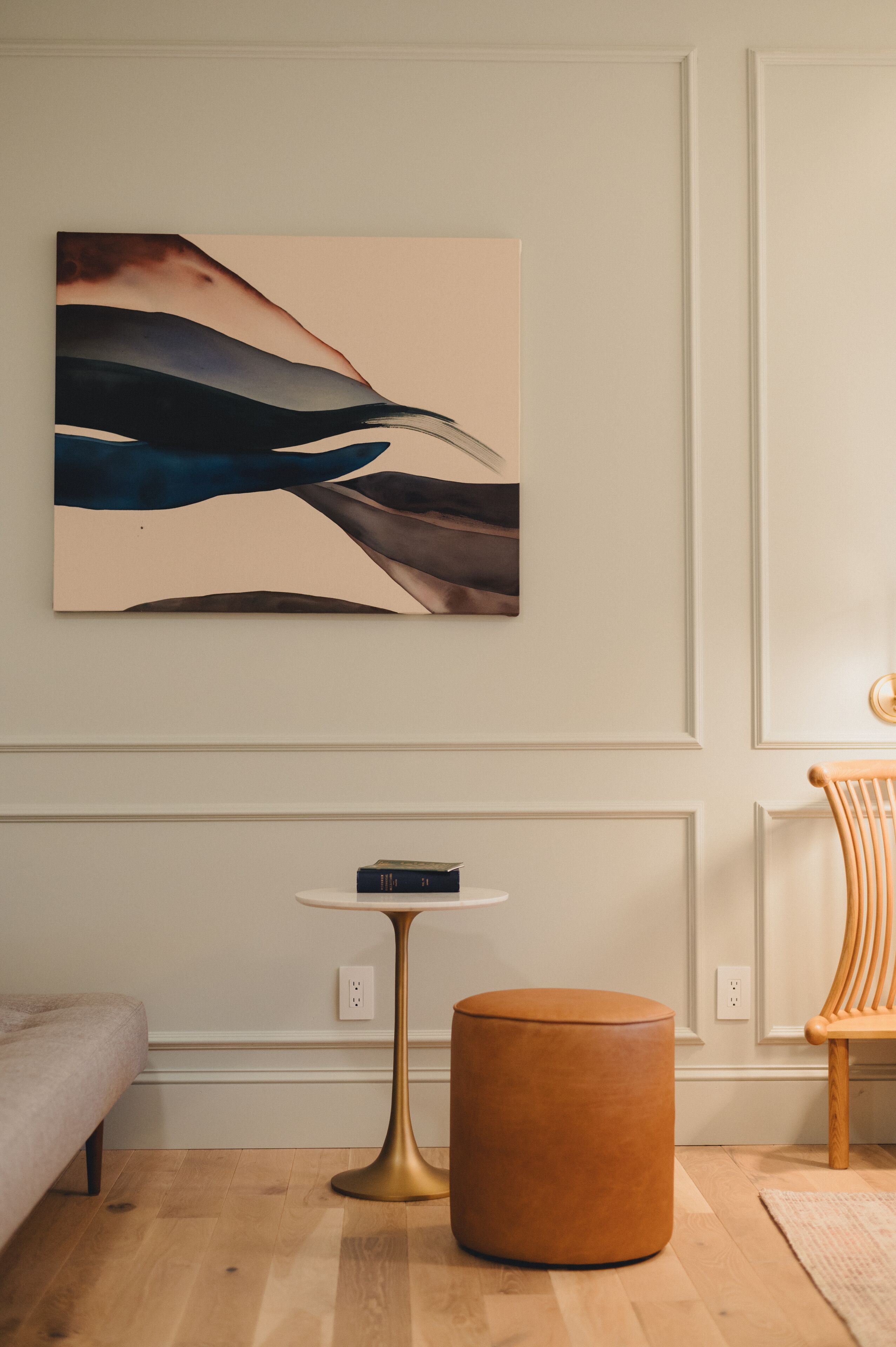 A room with a wooden floor, a round ottoman, a small table, and a wall-mounted abstract painting.