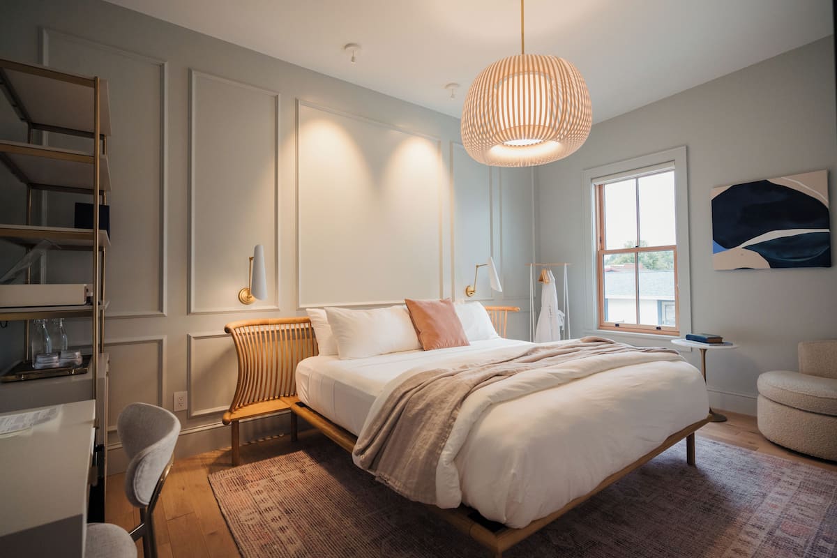 Un dormitorio moderno con una cama grande, una silla de madera, una mesita pequeña y una lámpara colgante.