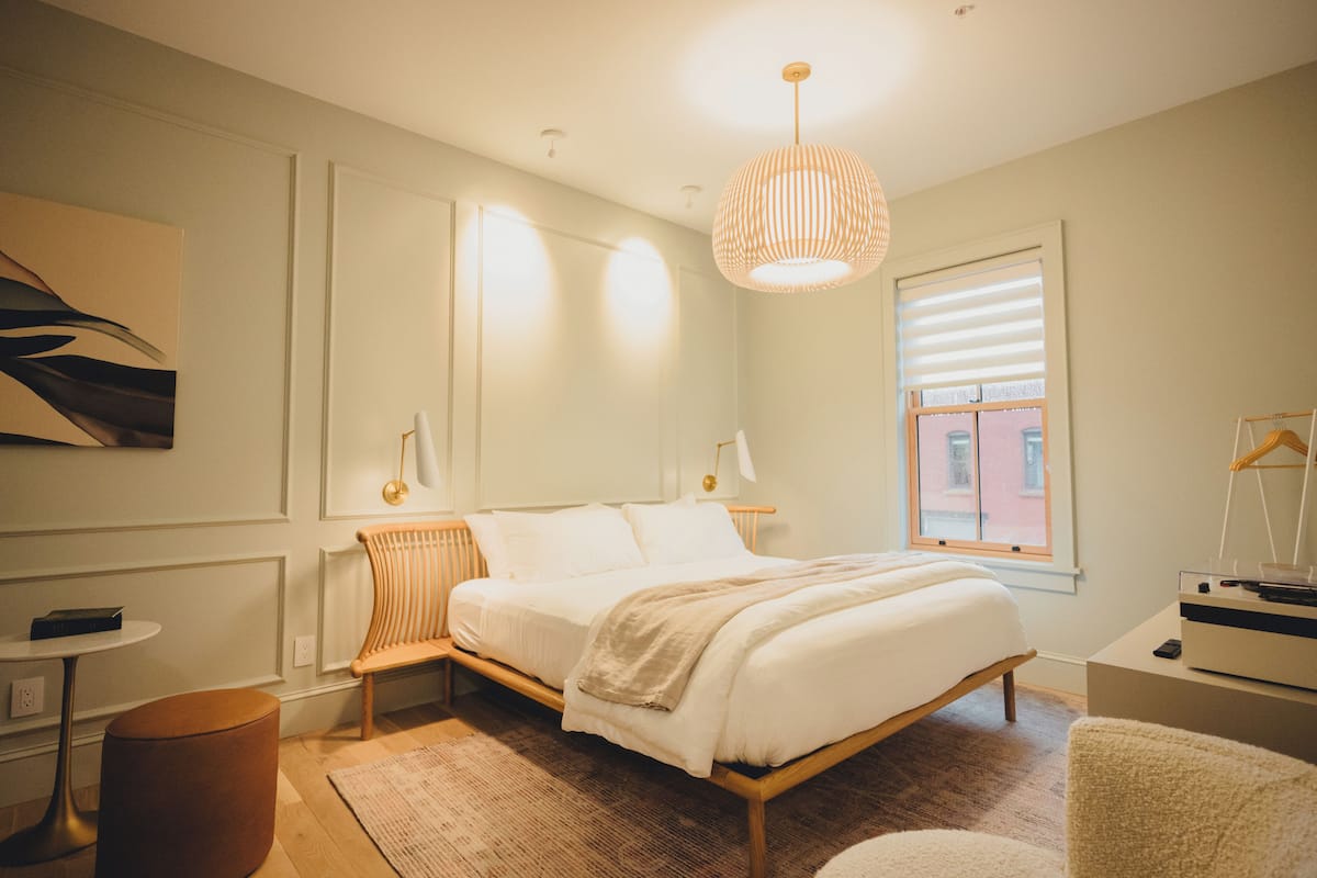 Un dormitorio moderno con una cama de madera, un banquillo redondo, una mesita y una lámpara colgante.