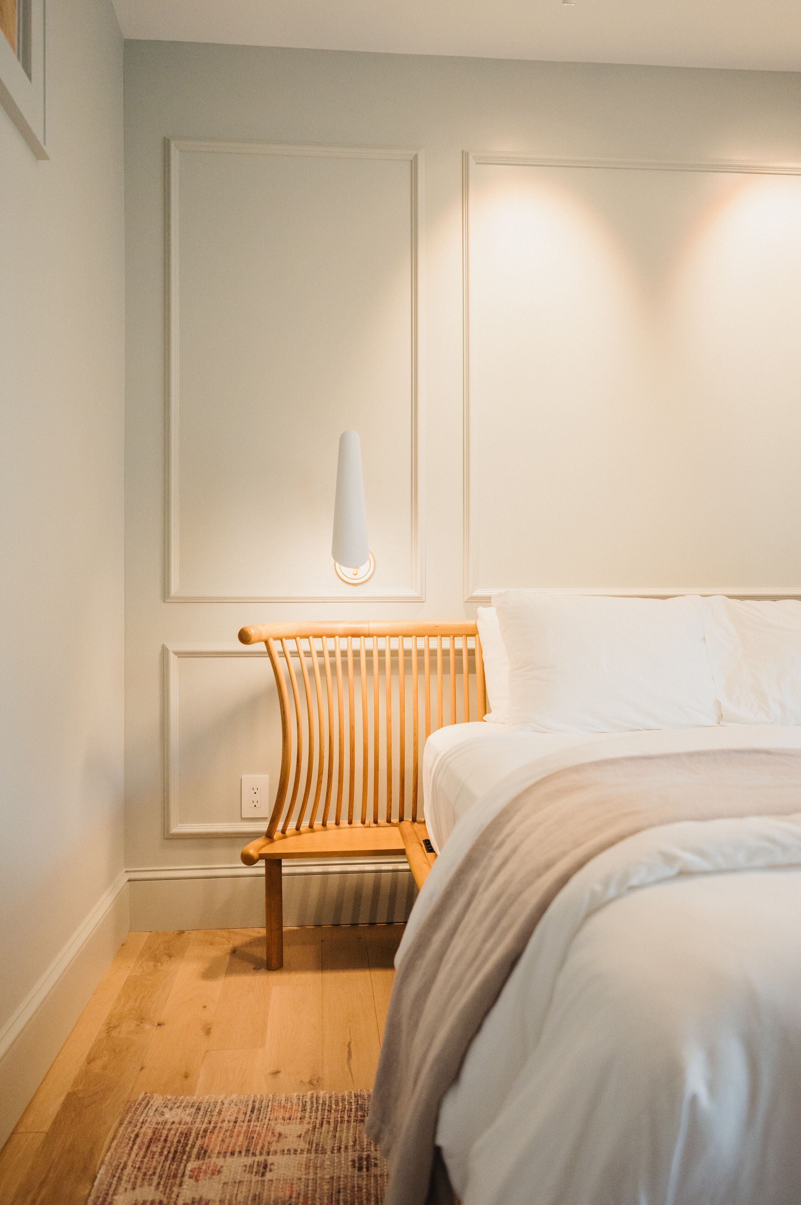 Une chambre avec une chaise en bois, un lit recouvert de linge de lit blanc et un luminaire fixé au mur.