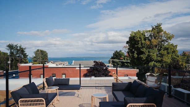A rooftop terrace with wicker furniture, a view of the sea, and a brick building in the background. - St. James Social (Bayfield)