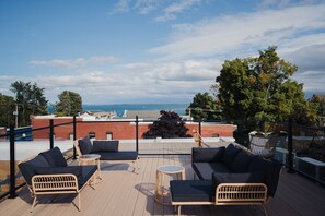 A rooftop terrace with wicker furniture, a view of the sea, and a brick building in the background. - St. James Social (Bayfield)