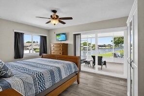 A bedroom with a bed, a ceiling fan, a TV, a wooden dresser, and a balcony with a view of a body of water. - Anchors Landing Waterfront Home | Dock | Foosball | Fire Pit | Close To The Beach | Dog Friendly (Palm Coast)
