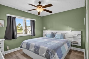 Ein Schlafzimmer mit Bett, Deckenventilator, Fenster mit Vorhängen, Nachttisch und Beistelltisch mit Lampe.