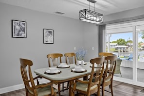 A dining area with a table set for four, wooden chairs, two framed artworks on the wall, and a view of a pool and outdoor area through a glass door.