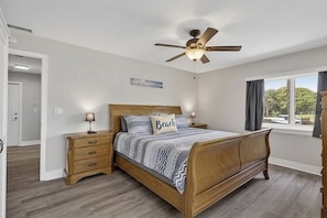 A bedroom with a wooden bed, two bedside tables, a ceiling fan, a window with curtains, and a door.