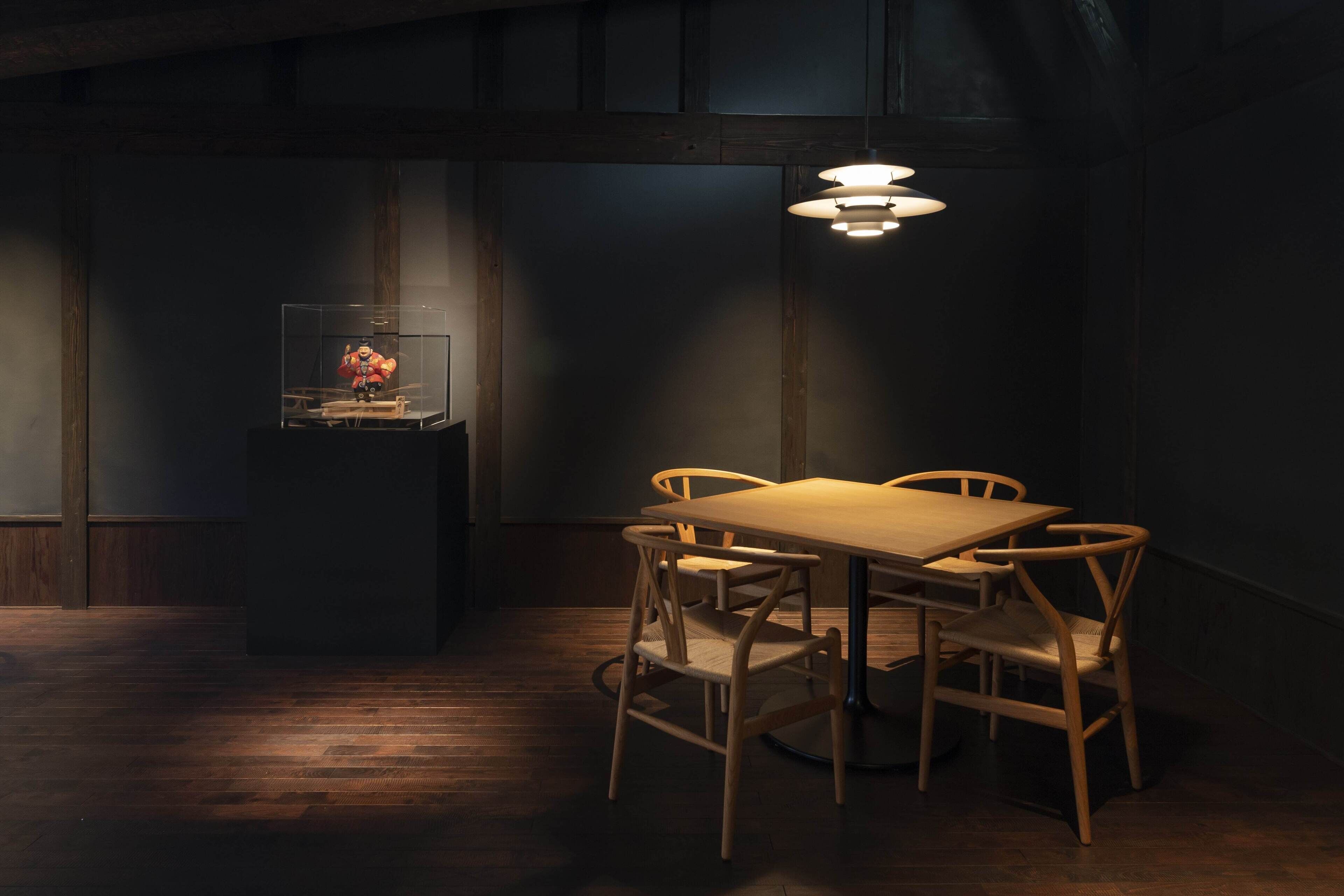 Um quarto com pouca luz, uma mesa de madeira com cadeiras, uma vitrine de vidro com uma miniatura e um lustre.