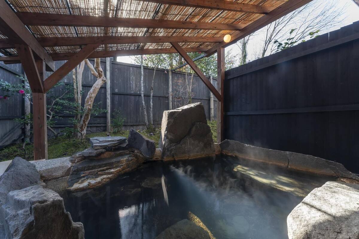 A hot spring area with a wooden pergola, stone seating, and a wooden fence in the background.