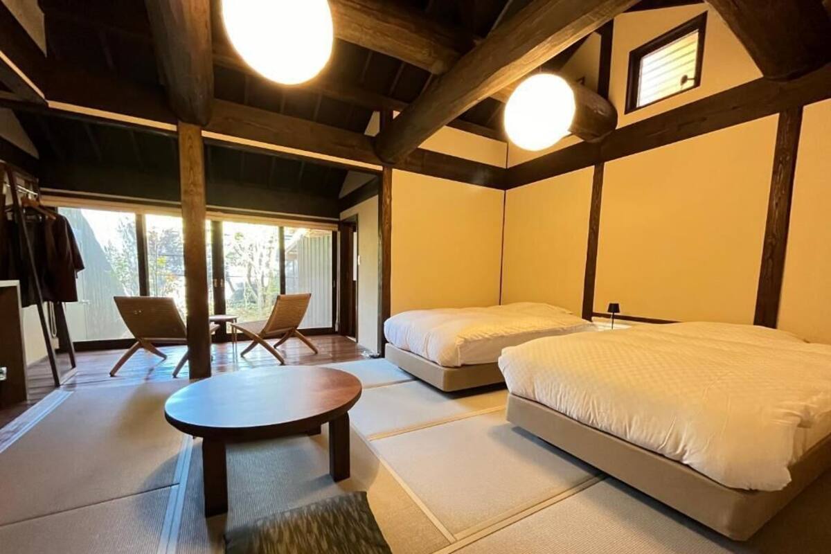 A room with two beds, a table, and chairs. There is a window with a view of trees.