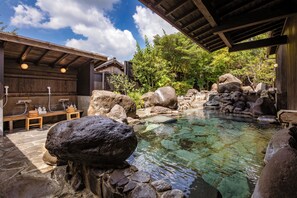 A traditional Japanese onsen with a wooden bathhouse, a clear hot spring, and surrounding rocks.