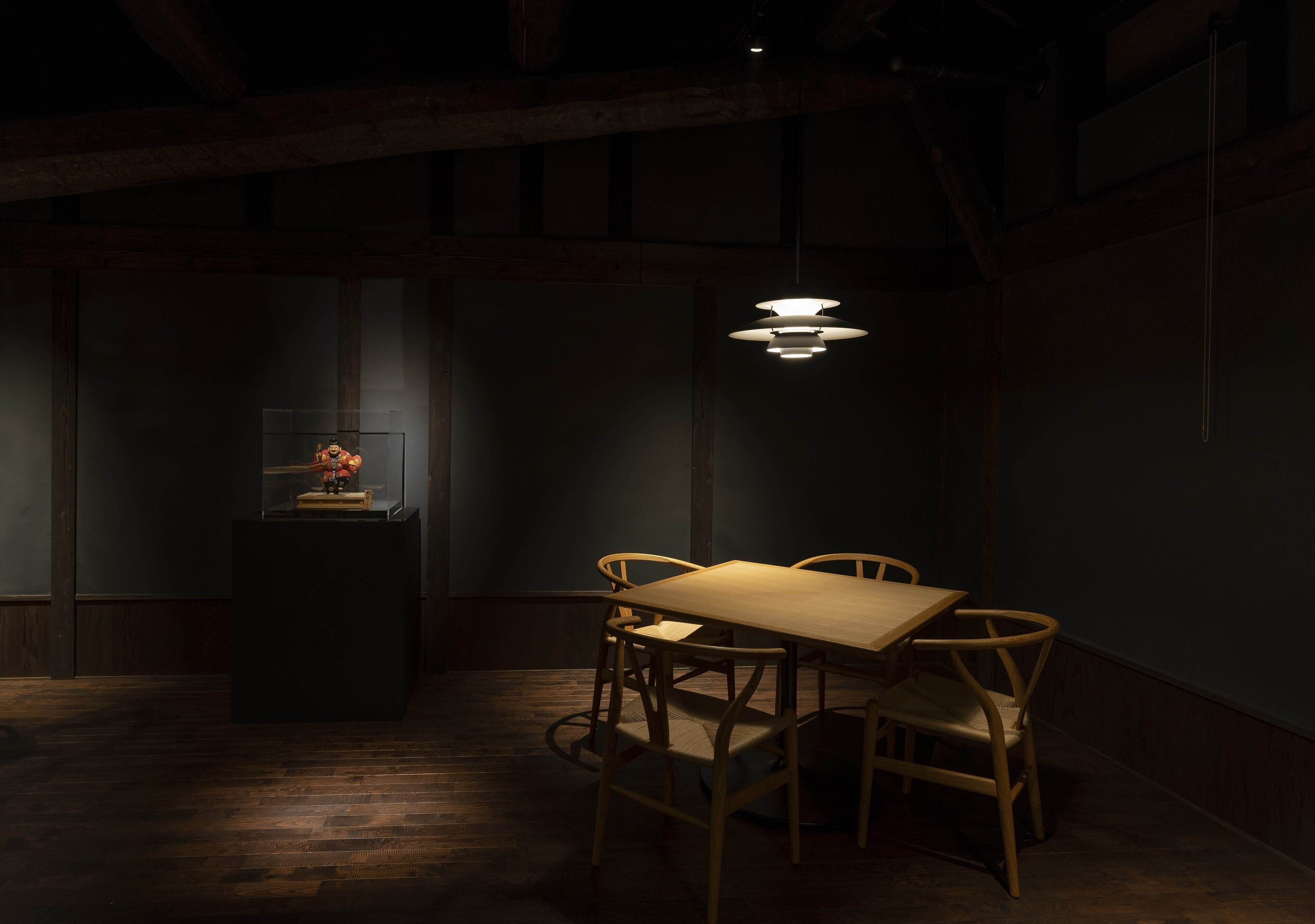 Et svagt oplyst rum med et træbord og stole, et lille piedestal med en pyntgenstand, samt en hængende lampe.