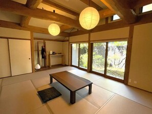 Una habitación tradicional japonesa con suelo de tatami, una mesa de madera baja, puertas correderas y grandes ventanales con vistas a la vegetación.