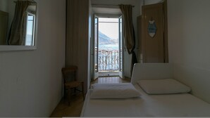 A bedroom with a bed, a chair, a window with curtains, a hanging light fixture, and a view of a mountain and sea through the window. - Casa Margherita in Varenna (Varenna)