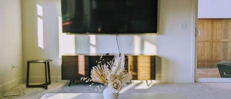 A modern living room with a flat-screen TV mounted on the wall, a white coffee table with a vase of dried flowers, and a small side table.