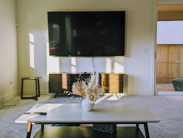 Ein modernes Wohnzimmer mit einem an der Wand montierten Flachbildfernseher, einem weißen Couchtisch mit einer Vase getrockneter Blumen und einem kleinen Beistelltisch.