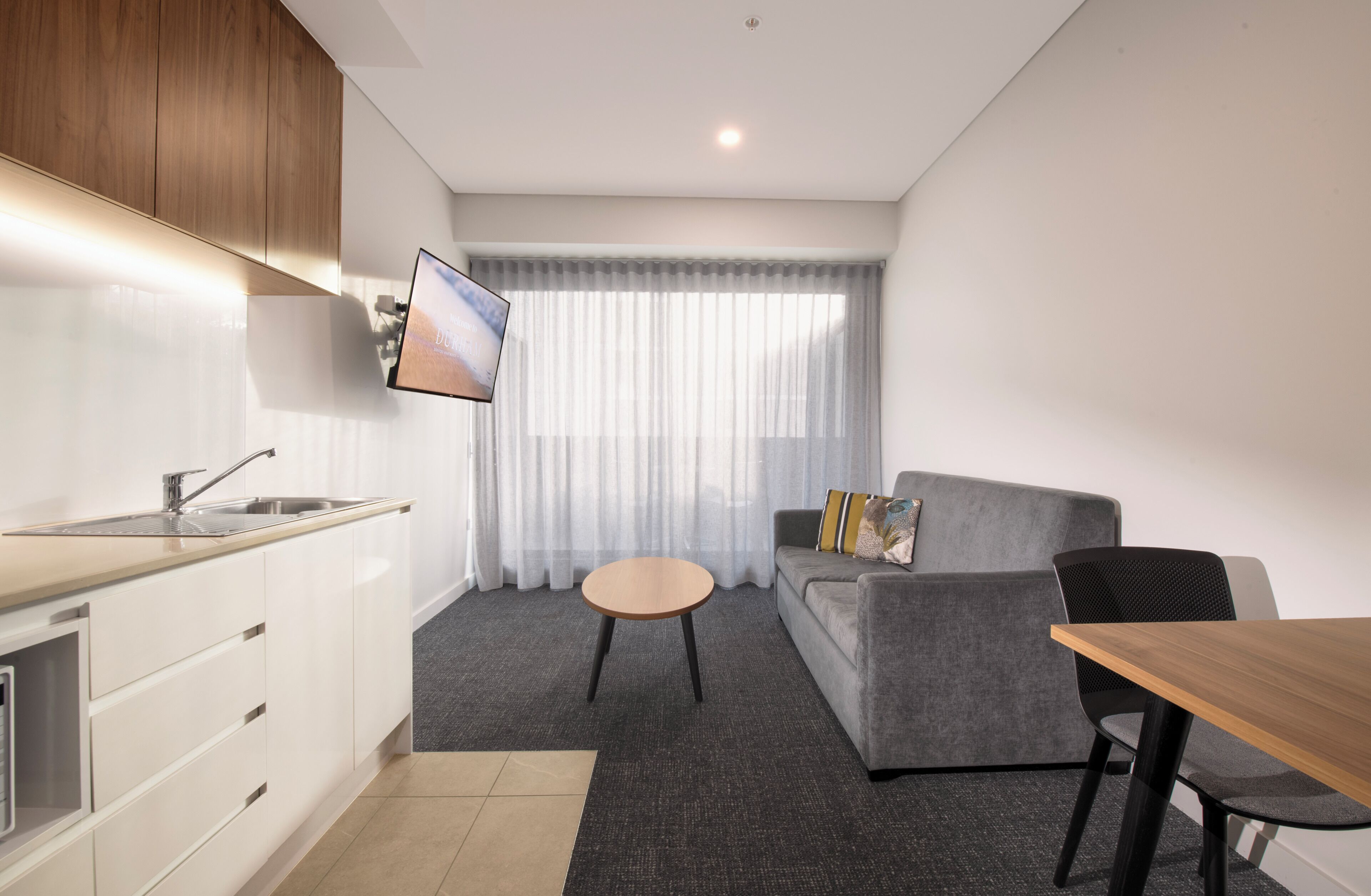 A modern kitchen with a sink and cabinets, a dining area with a table and chairs, a gray sofa with cushions, and a large window with sheer curtains.