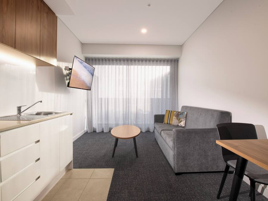 A modern kitchen with a sink and cabinets, a dining area with a table and chairs, a gray sofa with cushions, and a large window with sheer curtains.