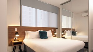 Habitación de hotel con una cama grande, dos mesitas de noche con lámparas, un espejo y una ventana con persianas.