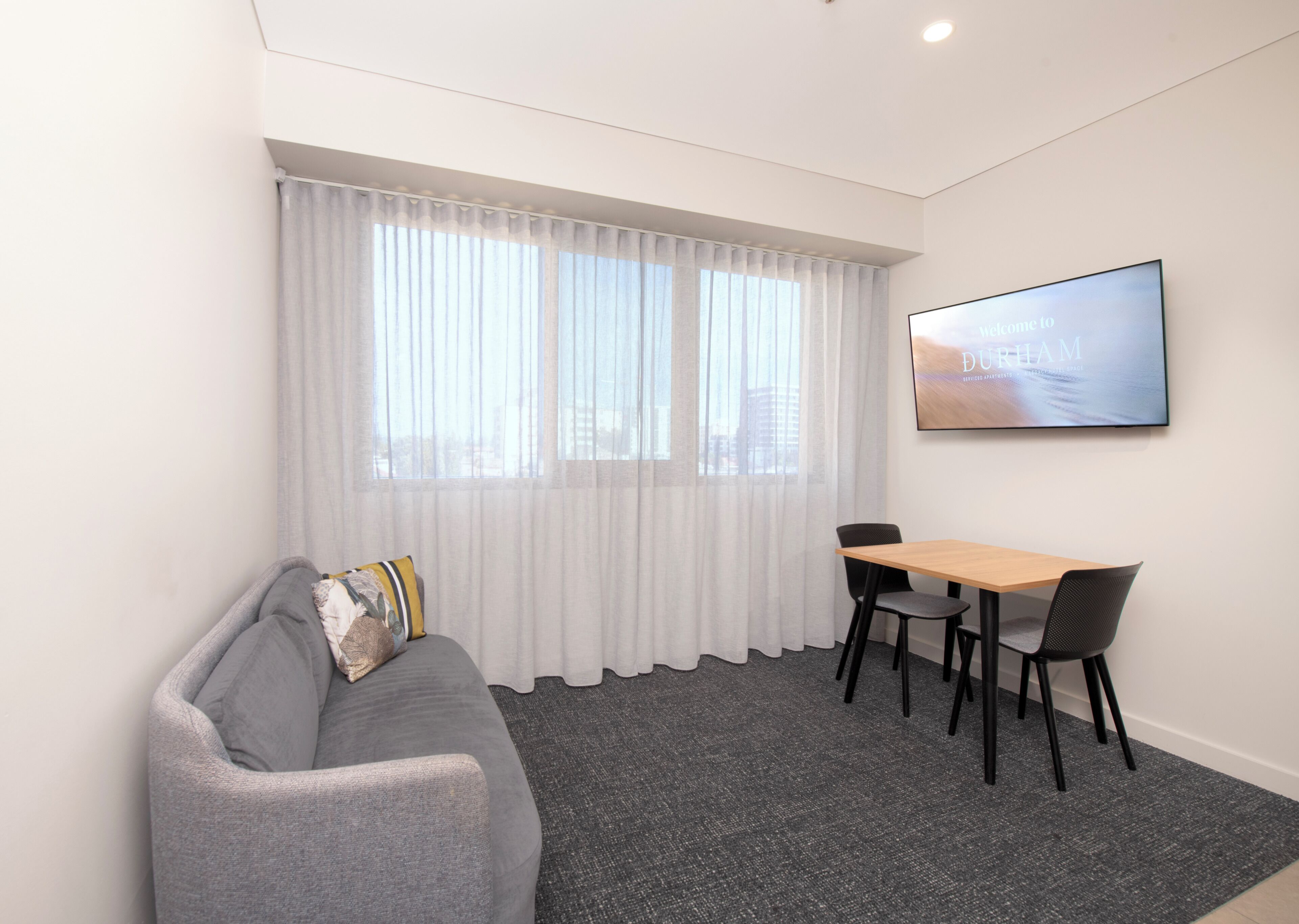 A modern room with a grey sofa, a wooden table, two black chairs, a large window with sheer curtains, and a framed welcome sign.