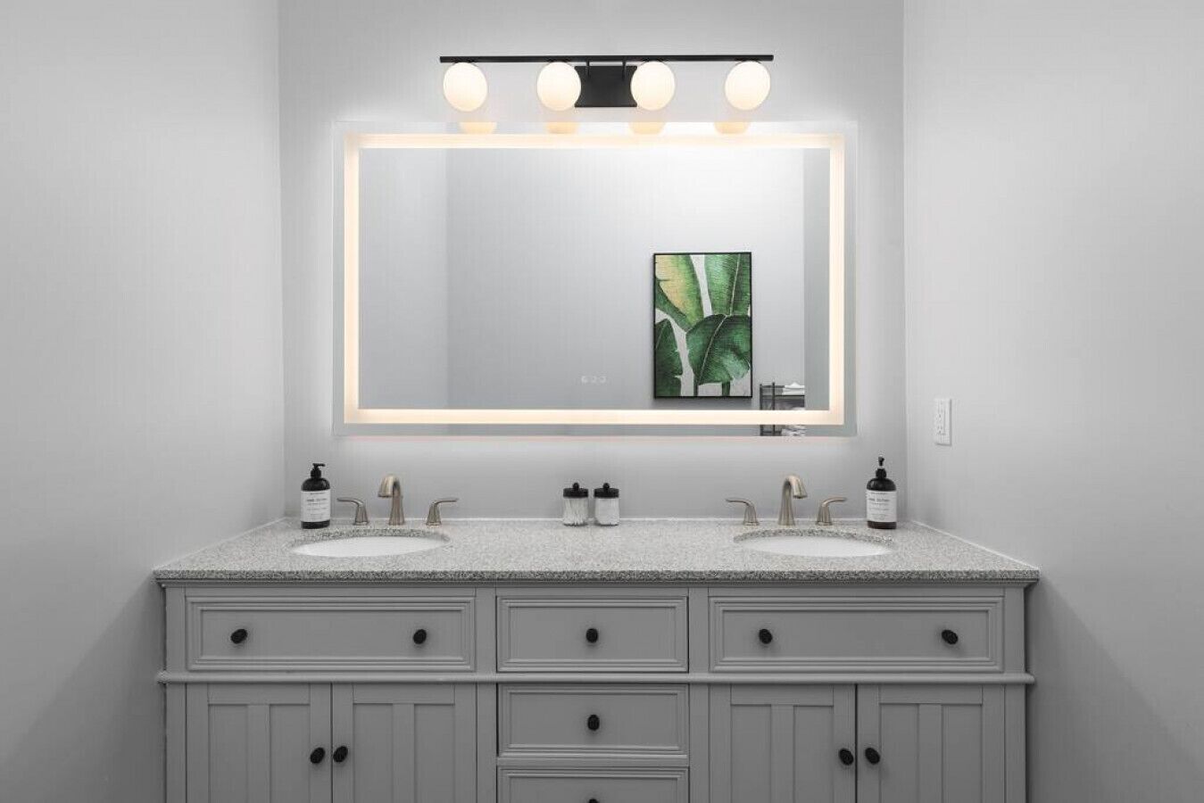 A bathroom with two sinks, a large mirror with lights, and a plant in the mirror.