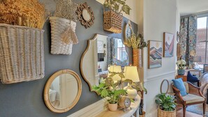 A room with a dark gray wall, a white console table with potted plants, wicker baskets, and a mirror. There are framed pictures and a blue chair with a cushion. - Against the Riverwalk/King (San Antonio)