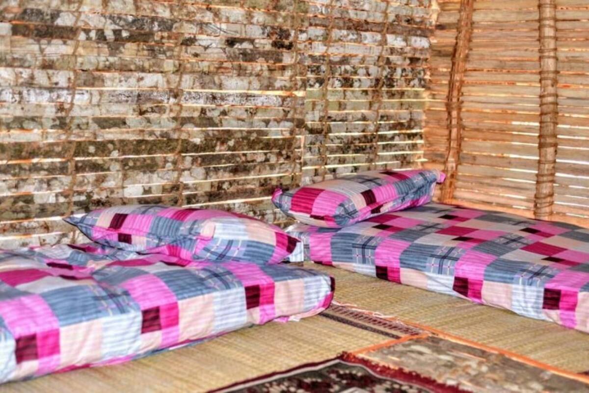 A room with a brick wall, two beds with patterned bedding, and a woven mat on the floor.