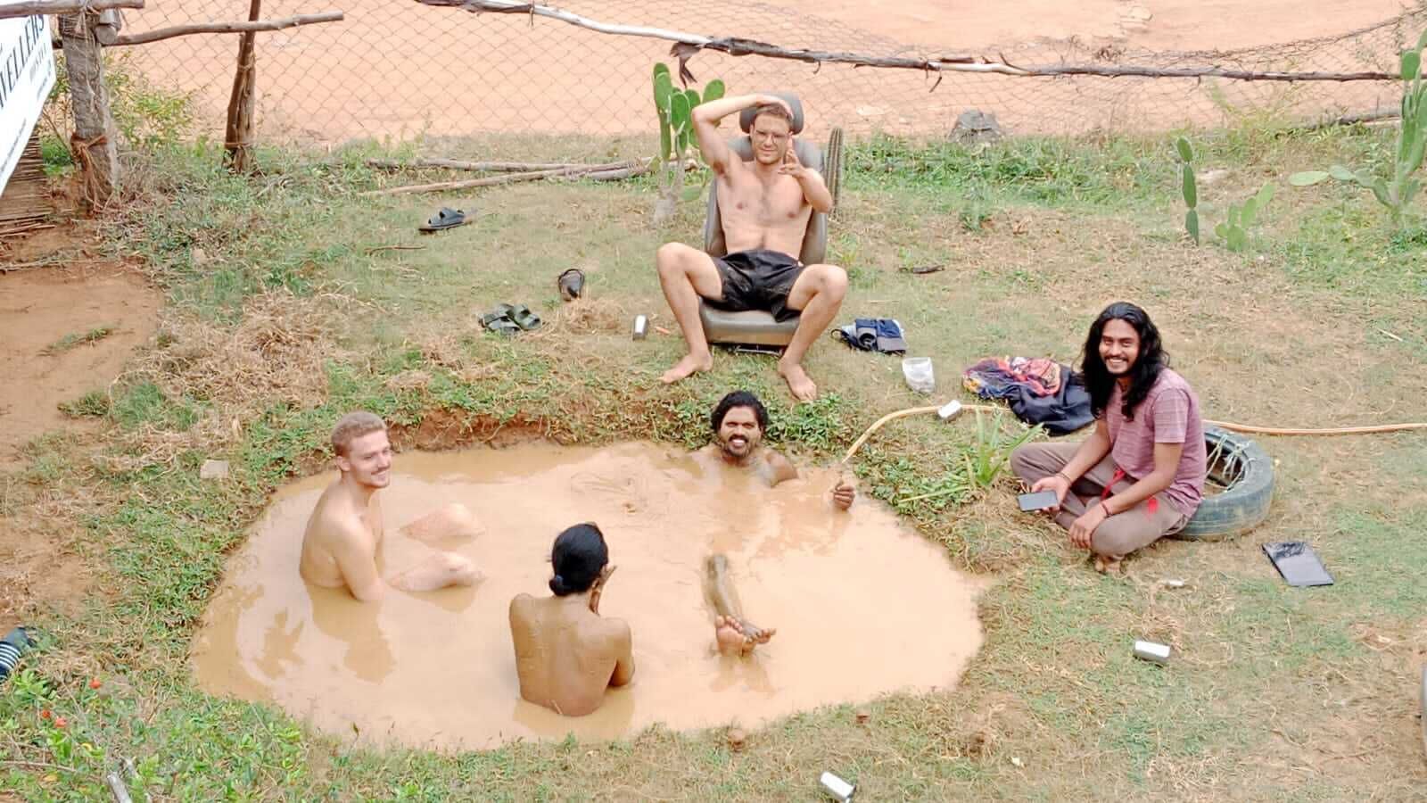 A group of people sitting around a small, muddy water-filled pit outdoors.