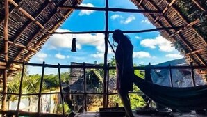 A person standing under a thatched roof structure, possibly a hut or a small building, with a view of the sky and distant landscape. - Time Travellers Hostel (Tindivanam)