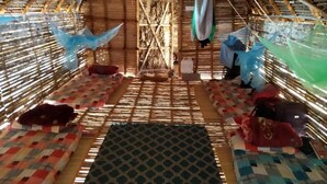 A room with a thatched roof, bamboo walls, and a central woven partition. There are mats on the floor and hanging laundry. - Time Travellers Hostel (Tindivanam)