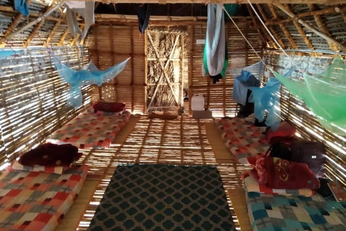A room with a thatched roof, bamboo walls, and a central woven partition. There are mats on the floor and hanging laundry.