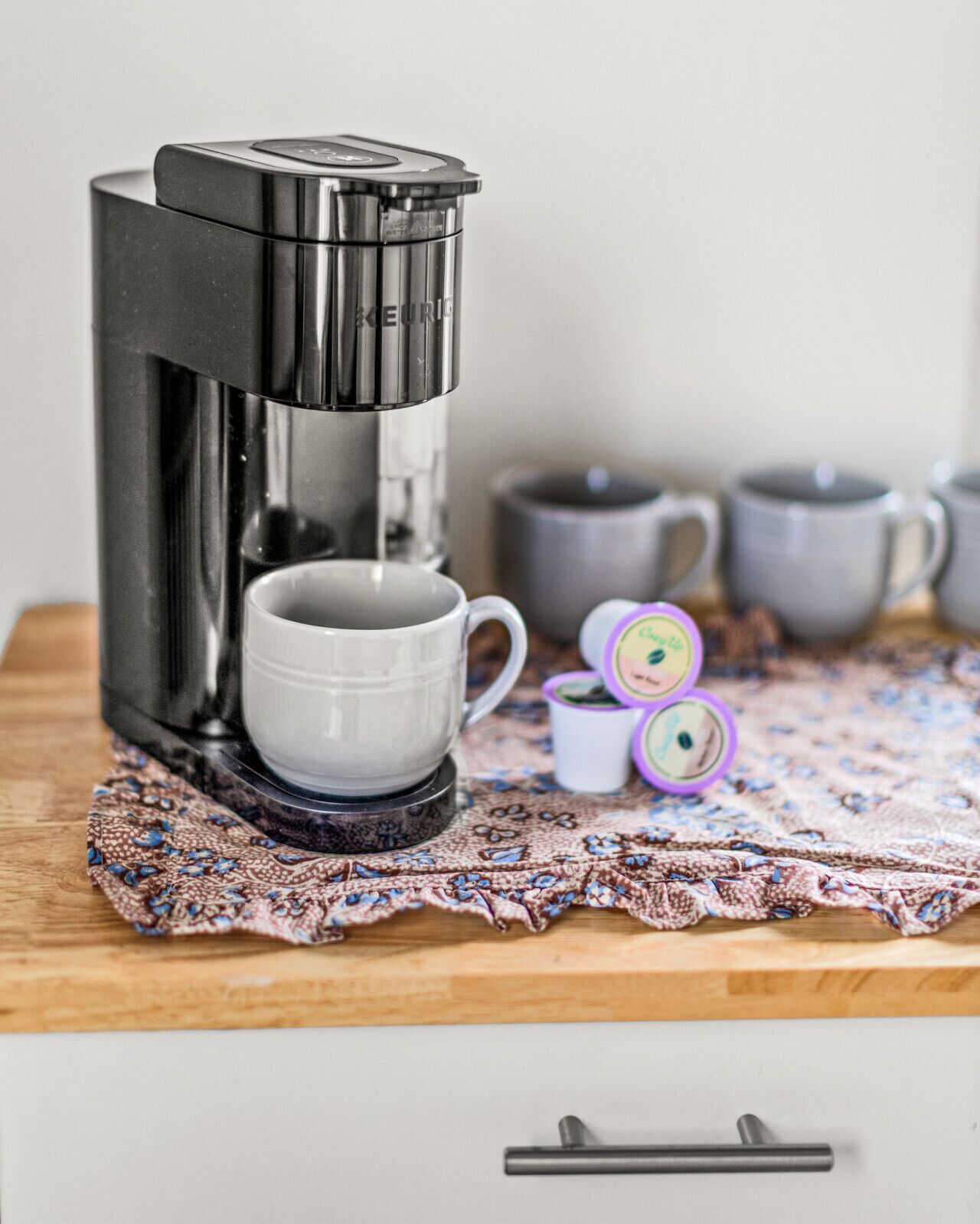 A coffee maker on a wooden table with a lace tablecloth, a white mug, and two small containers.