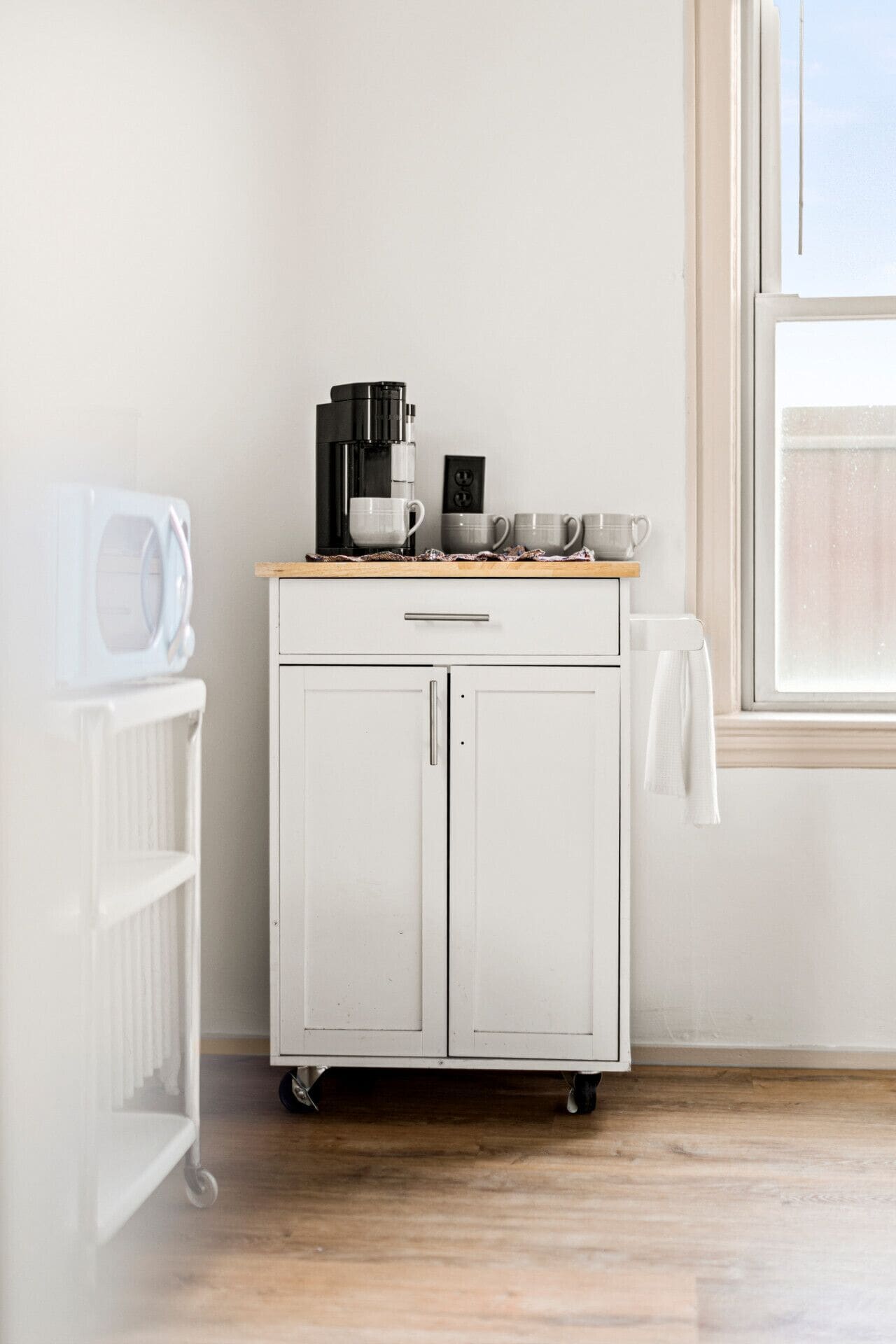 A white kitchen with a microwave, coffee maker, and mugs on a wooden countertop.
