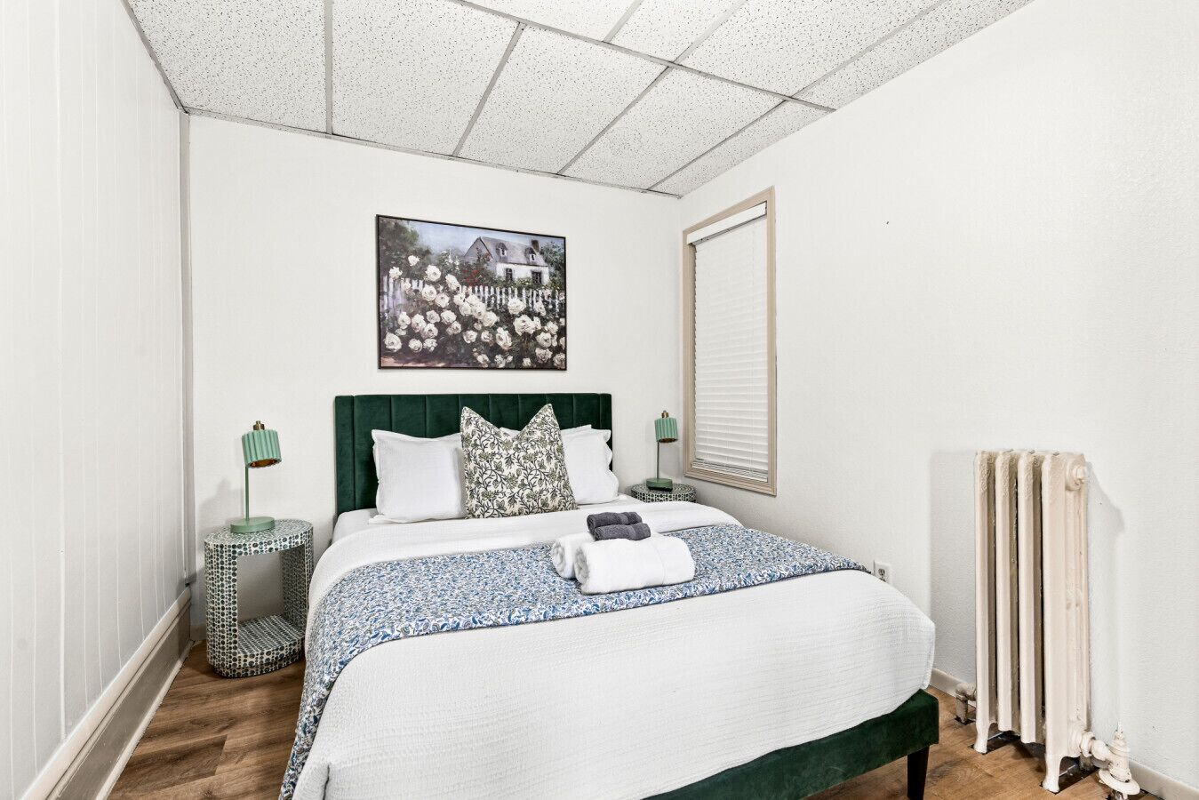 A bedroom with a bed, a radiator, a picture on the wall, and a window with blinds.
