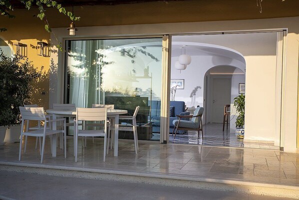 Un patio al aire libre con una mesa y sillas, un ventanal de vidrio que da a un espacio interior y una planta en maceta.
