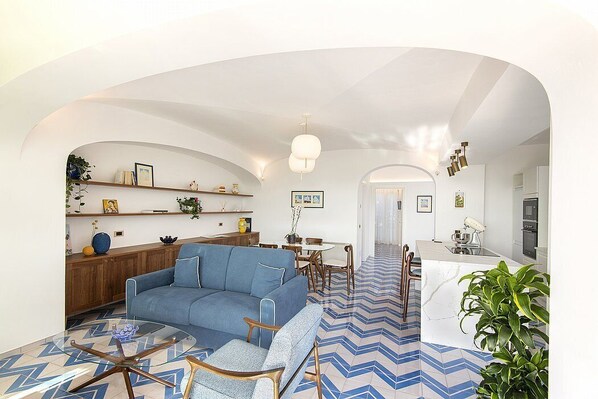 A modern living room with a blue sofa, a glass coffee table, and a dining area with a white island.