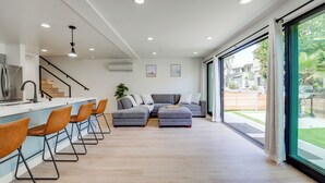 Een moderne woonkamer met een grijze modulaire bank, een keukenhoek met barstoelen en een grote schuifpui naar een tuin.